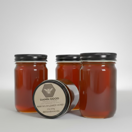 Jars of raw wildflower honey, with one labeled 'Damn Good Raw Wildflower Honey 1 lb', on a wooden surface.