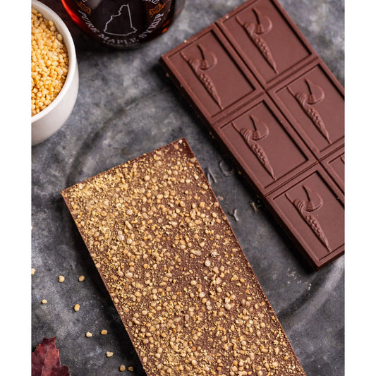 Two chocolate bars on a gray surface with a bowl of seeds and a bottle in the background.